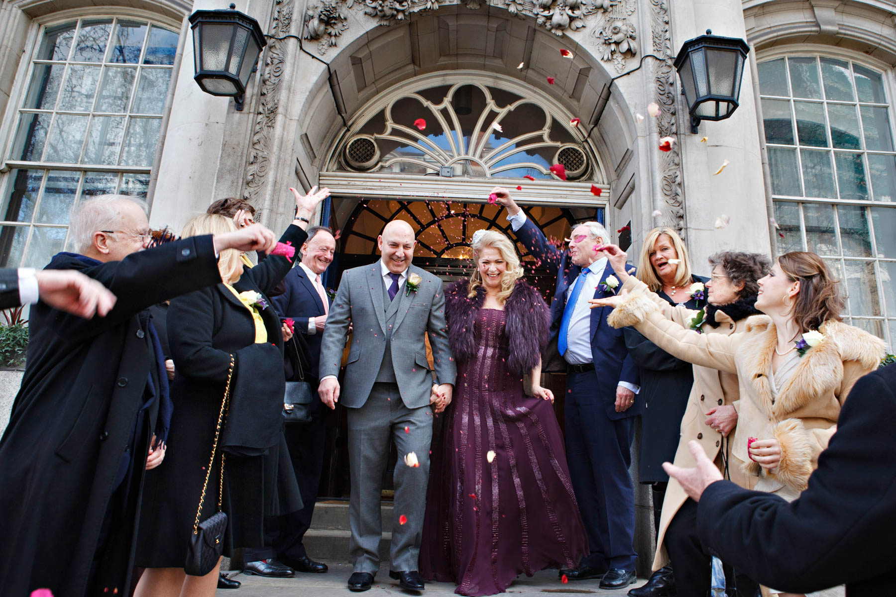 A mature bride and groom exit their wedding ceremony into lots of confetti thrown by their guests on the steps of Chelsea Old Town Hall. The bride is wearing a dark purple wedding dress.