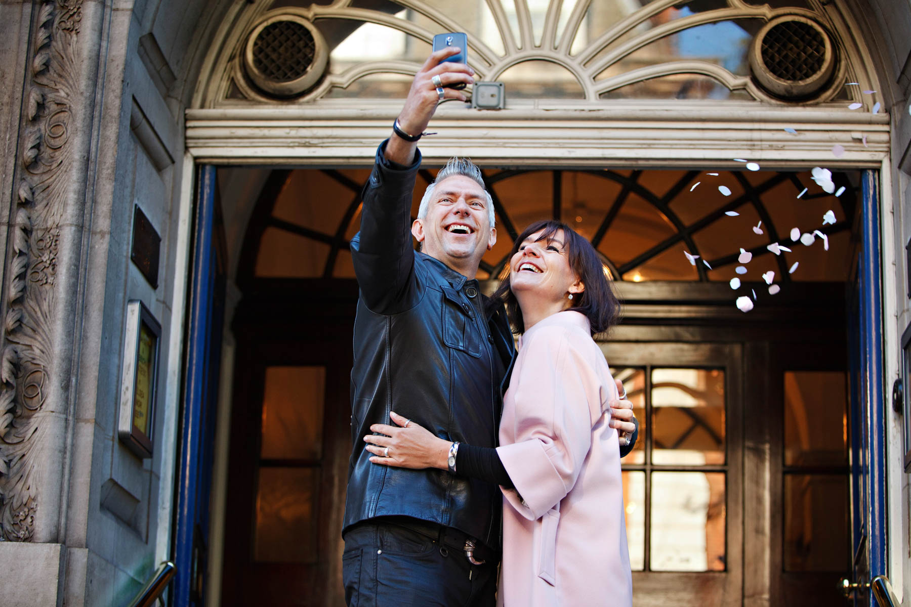 A bride in pink and groom in black leather jacket take selfies as they throw confetti at themselves after their elopement wedding ceremony in the Rossetti Room.