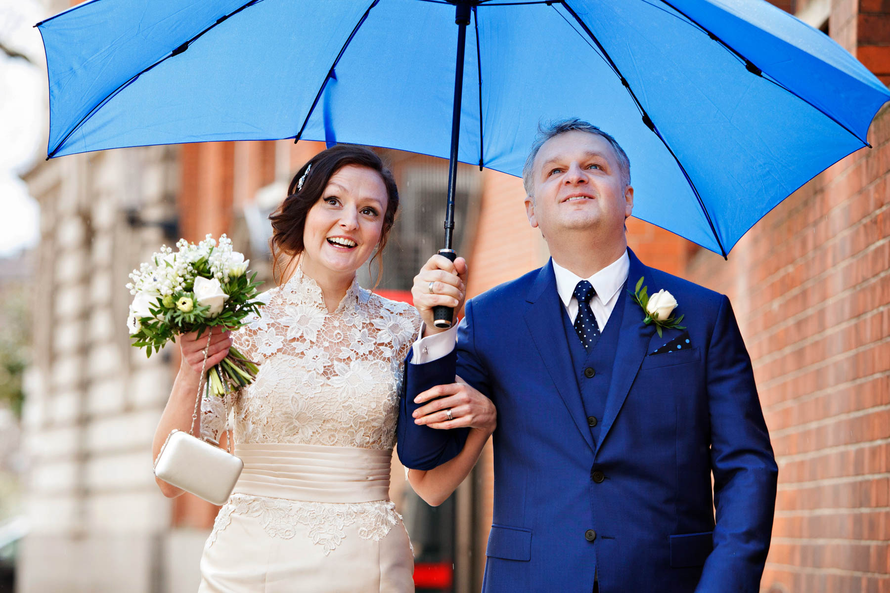 A bride and groom carrying a blue umbrella look up at the sky as it rains on their wedding day. But they are not letting the wether spoil their fun or detract from the joy of their Chelsea Register Office wedding.
