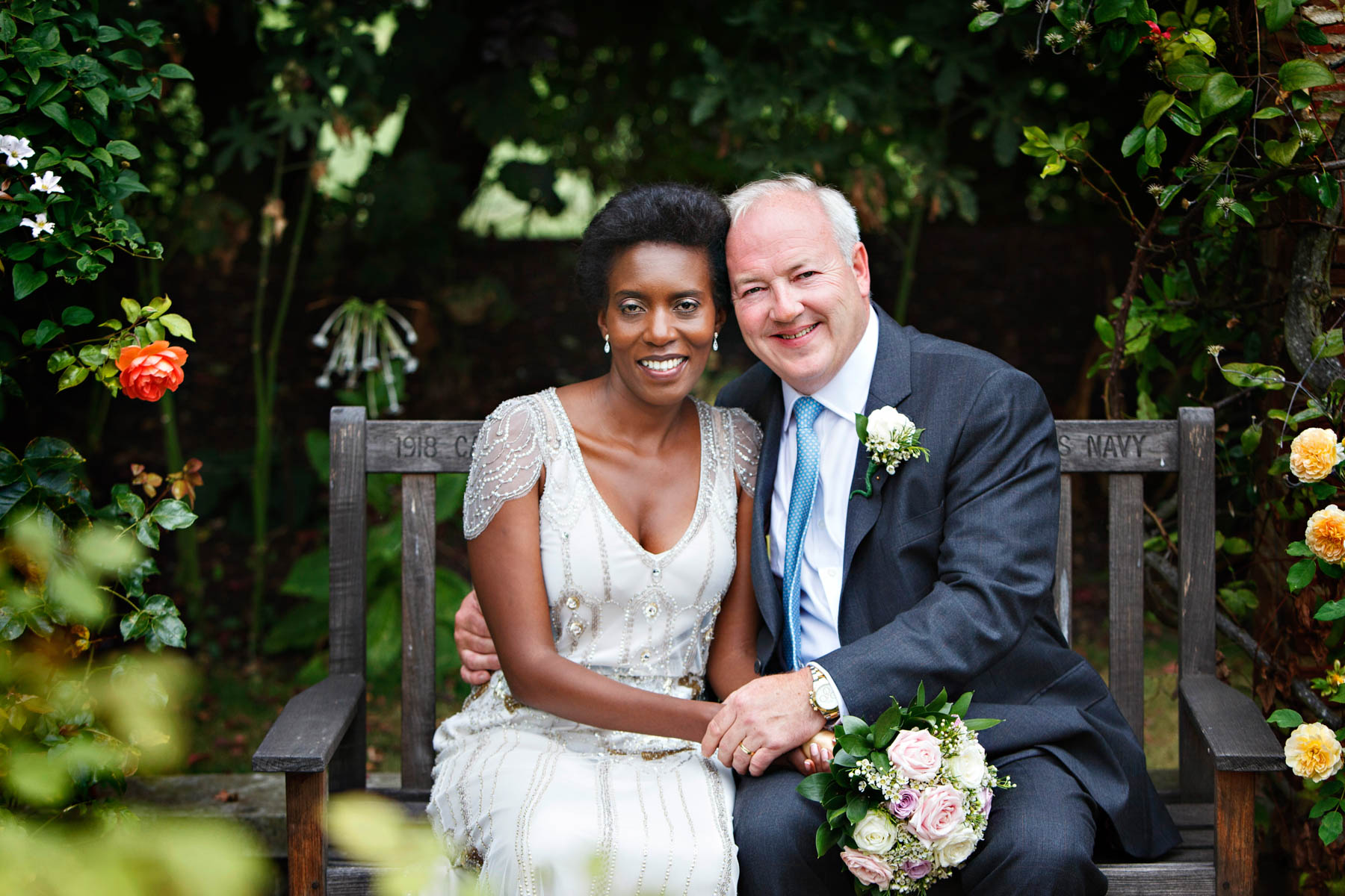 A bride and groom celebrate their Chelsea Old Town Hall Wedding at the super famous Hurlingham Club. They are photographed during their wedding portrait session, sitting on a bench in the landscaped gardens of the Hurlingham Club.