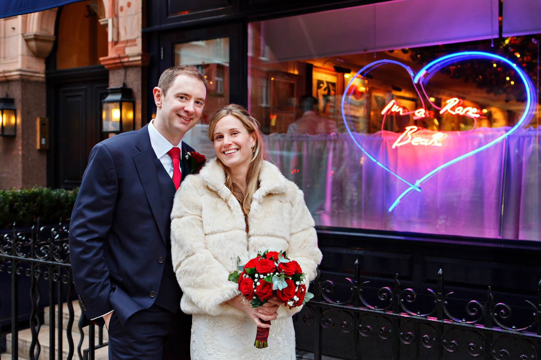 Winter wedding portrait of a bride and groom wrapped up warm, in front of a restaurant window with a big pink flourescent heart in it. The bride is wearing a white faux fur jacket and carrying a red rose bouquet.