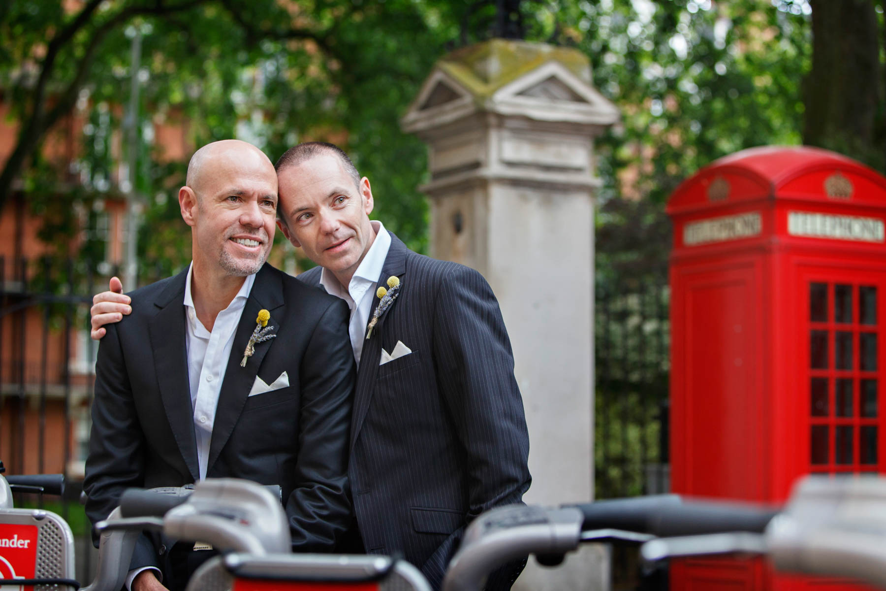 Two grooms snuggle up to each other after their same-sex civil partnership ceremony at Mayfair Library. A classic red London phone booth can be seen in the background.