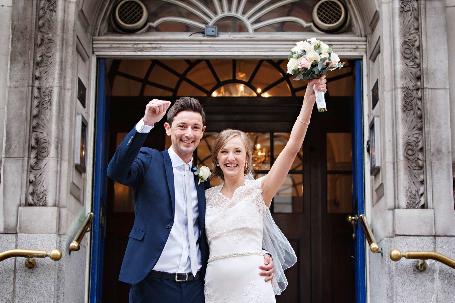 A bride and groom wave their arms in the air cheering to celebrate getting married at Chelsea Old Town Hall.
