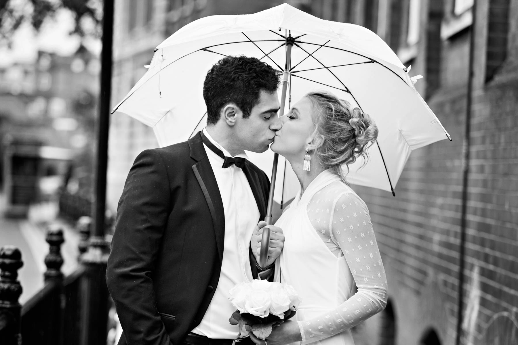 A black and white image of a bride and groom kissing under a white umbrella.