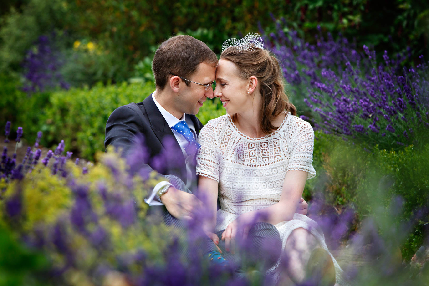 A pair of newlyweds sit in the gardens of Morden Park House, amongst lots of beautiful flowering lavender. They are sharing an intimate moment, with their foreheads resting against each other.