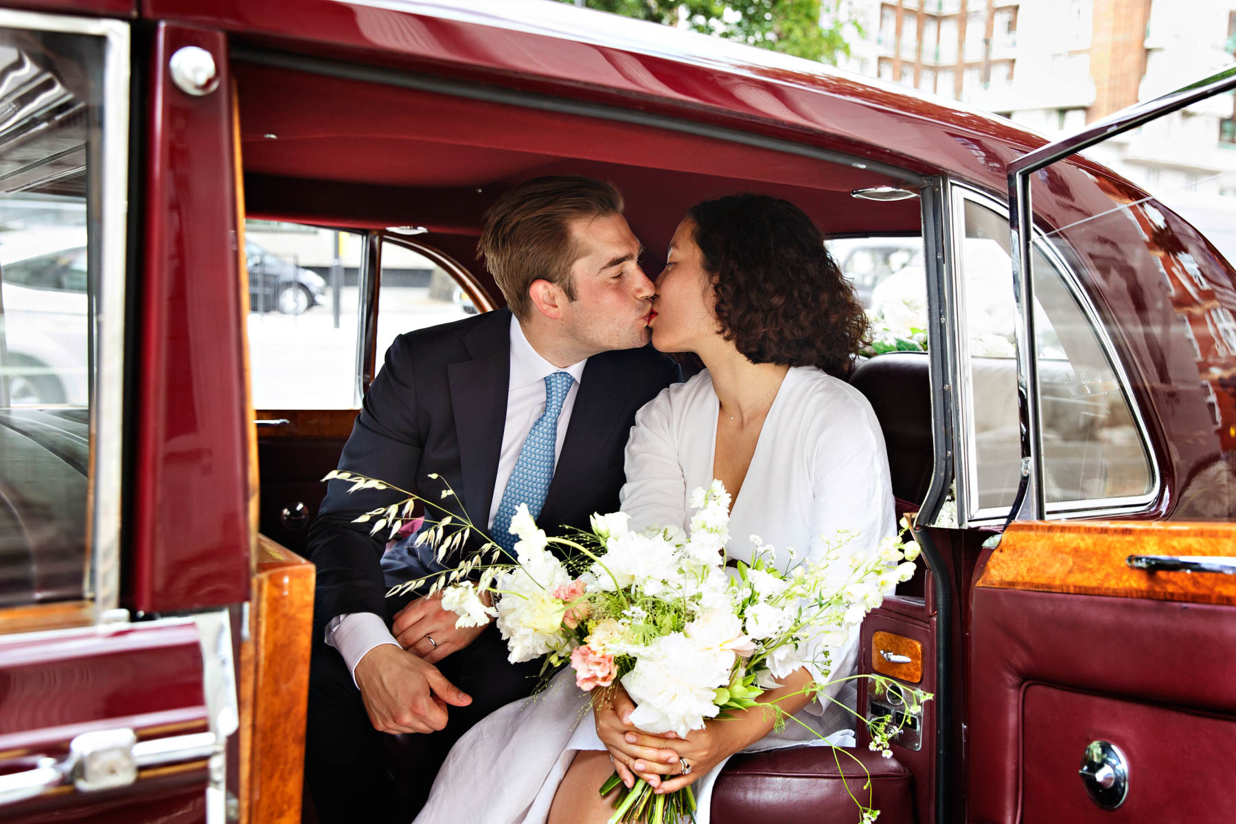 A bride and groom kiss in the back of a maroon vintage Rolls Royce, after their intimate wedding ceremony in the Marylebone Room.