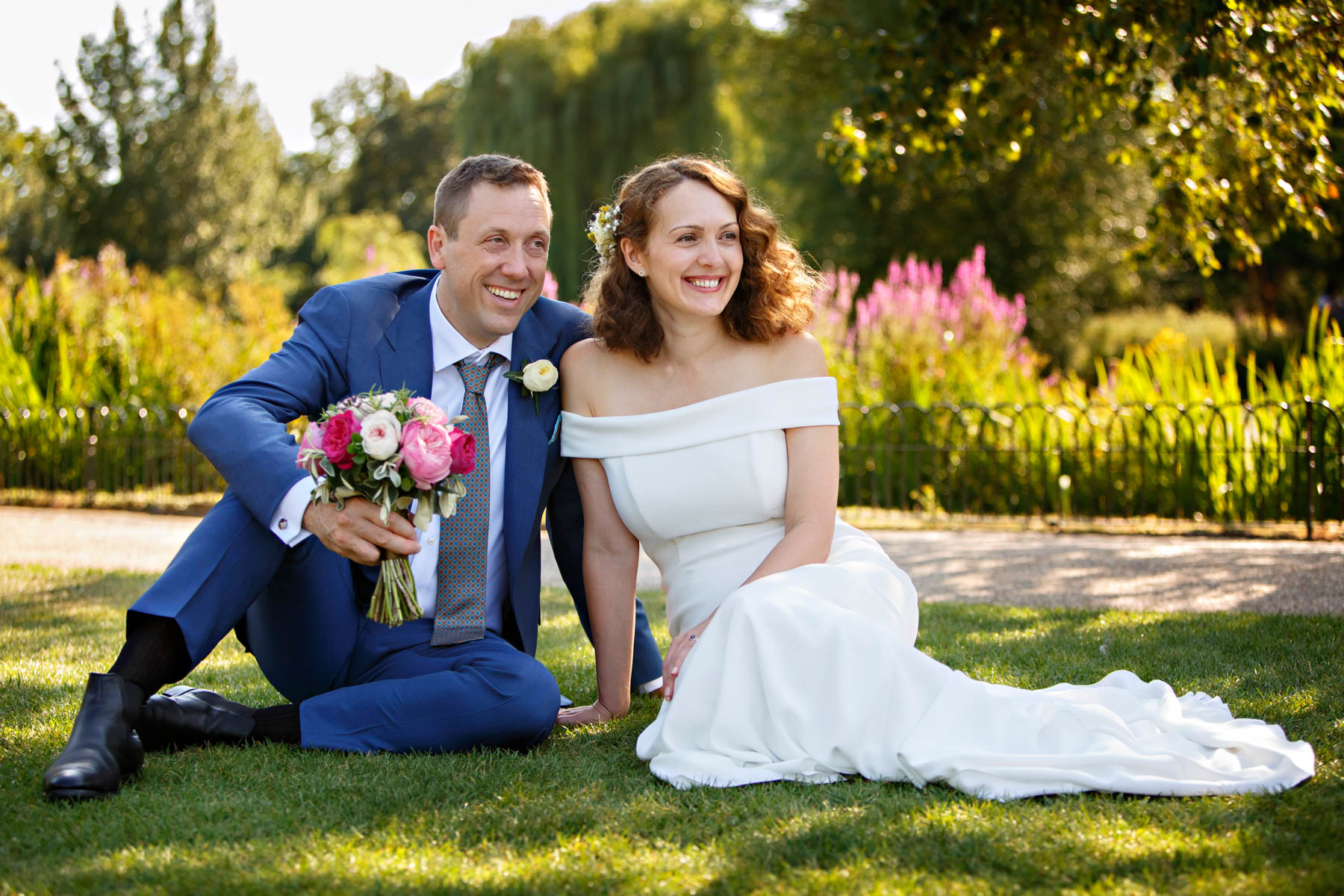 A bride and groom sit on the grass in the rose garden of Regent's Park, after their Soho Room Wedding at Old Marylebone Town Hall. The groom is holding the bride's bouquet and they're both looking off into the distance, smiling.