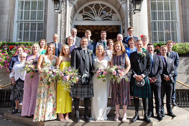 A bride, groom and their wedding guests stand on the steps of Chelsea Old Town Hall for a group wedding photo.