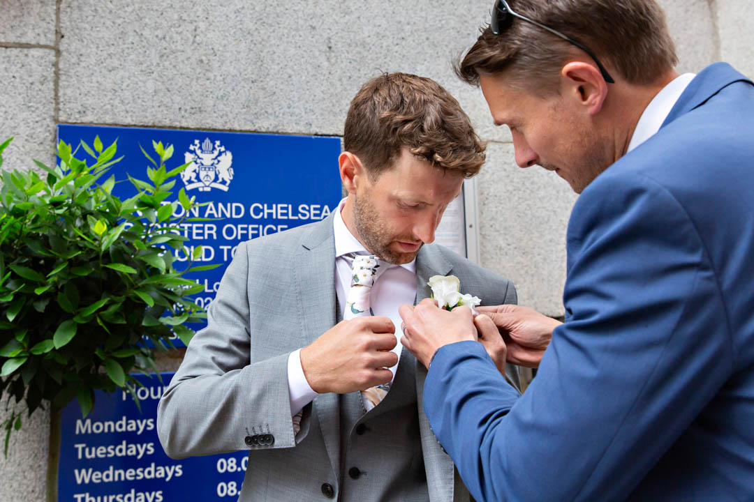The best man helps the groom attach his button hole outside Chelsea Old Town Hall.