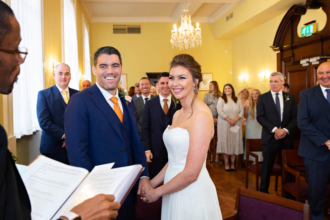 The bride and groom smile at the registrar whilst exchanging their vows in the Brydon Room at Chelsea Old Town Hall
