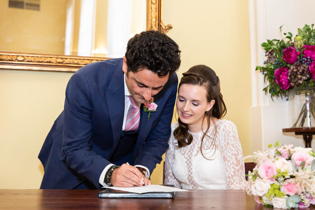 The groom signs the wedding schedule as his bride looks on in the Brydon Room at Chelsea Old Town Hall.