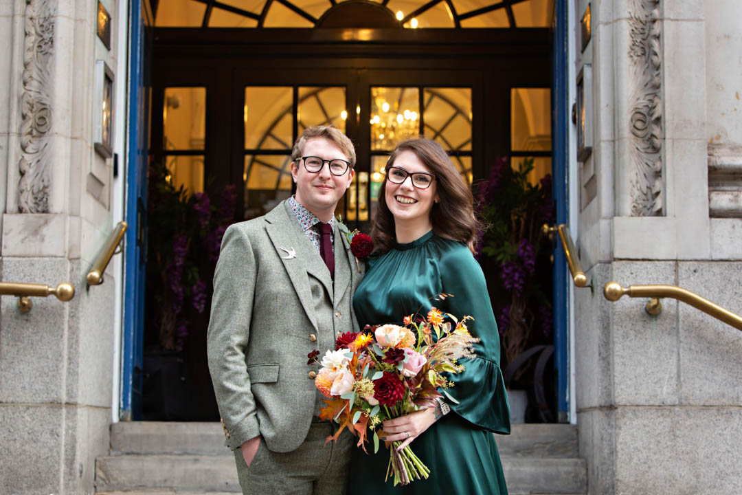 An autumn feel wedding portrait of a smiley wedding couple on the steps of Chelsea Old Town Hall. The bride is wearing a green sating wedding dress and is holding a large bouquet in autumn rust, orange and red colours.