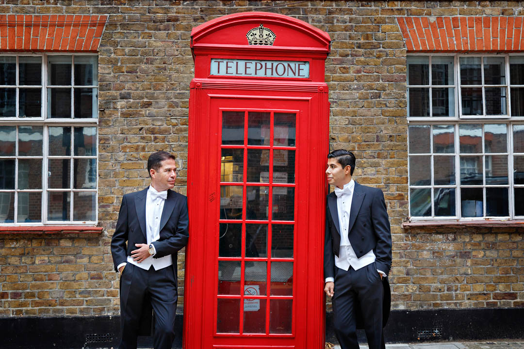 Two elegant grooms pose with red London phone box after their Brydon Room civil wedding at Chelsea Old Town Hall