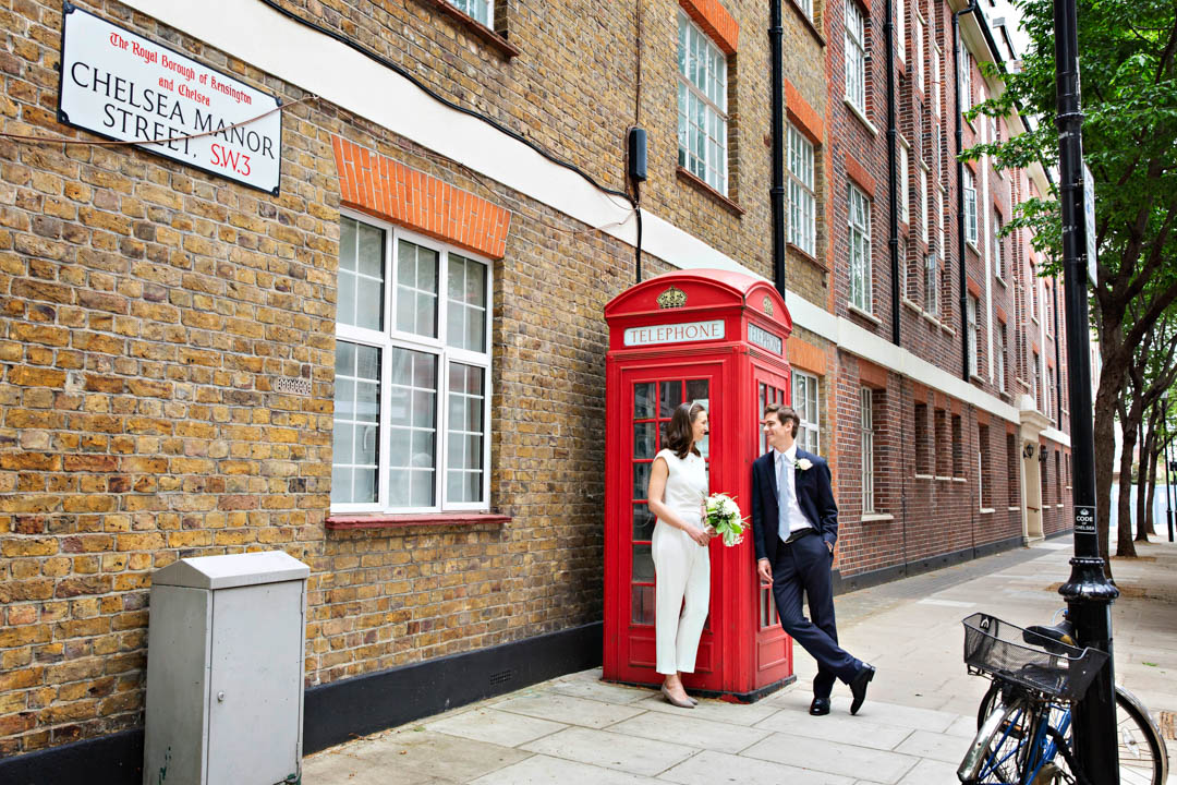 A bride and groom pose for their relaxed wedding portraits, leaning against a red London phone box with a Chelsea Manor Street sign visible in the shot.