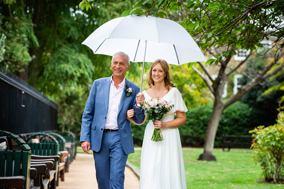 A bride and groom walk in St. Luke's garden's in Chelsea under a white wedding umbrella, after their Brydon Room wedding in Chelsea Old Town Hall