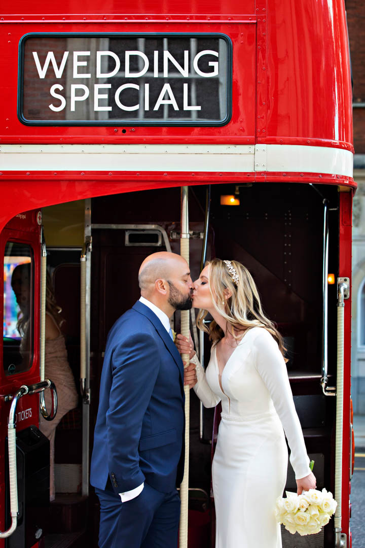 A bride and groom kiss on the back platform of their vintage red London routemaster bus outside after their Brydon Room wedding ceremony.