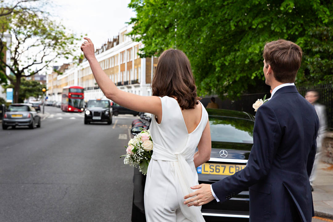 Bride in a white jumpsuit hails a black London taxi with her groom after their Chelsea Old Town Hall Wedding Ceremony