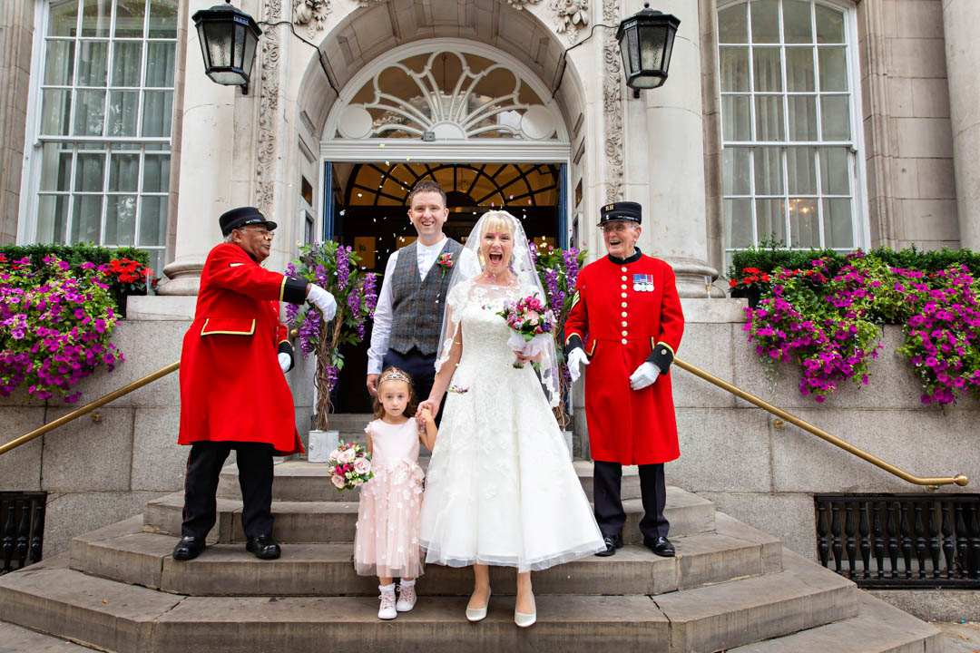 Chelsea Pensioners throw confetti over bride and groom who are accompanied by their little daughter.