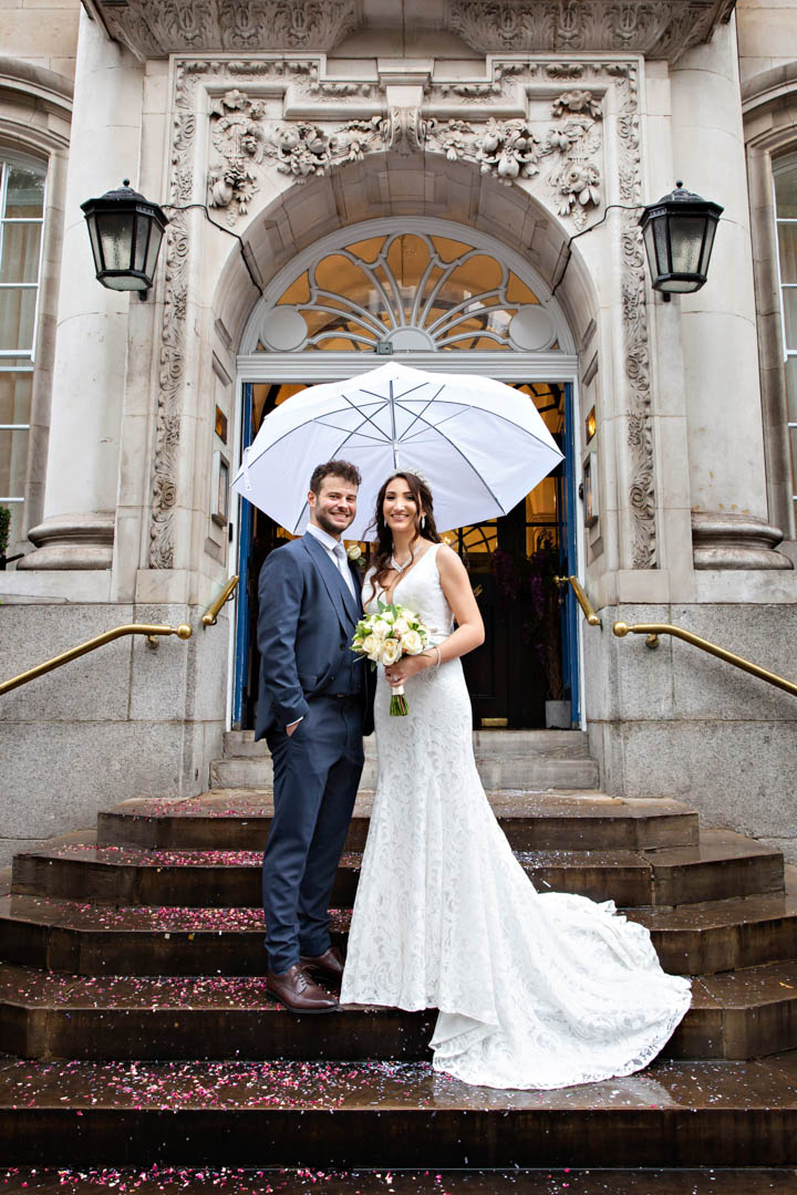 Elegant bride and groom cuddle up under a big umbrella on the steps of Chelsea Town Hall after their intimate civil wedding.