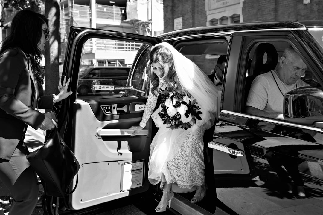 A bride wearing full-length white lace and a cathedral-length veil steps out of a London black cab, carrying a large white and deep maroon wedding bouquet.