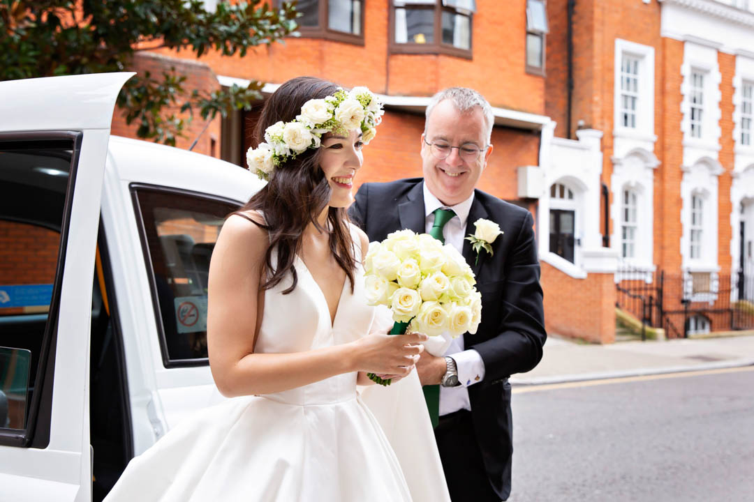 A smiling bride wearing a floral crown emerges from a white taxi while her father in a dark suit and emerald tie watches.