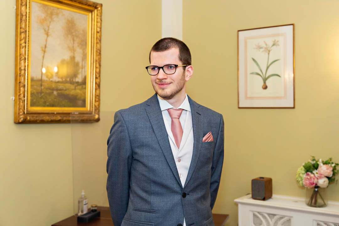 A groom in a grey jacket with a pink tie and pocket square waits for his bride to enter the Rossetti Room for their civil marriage.
