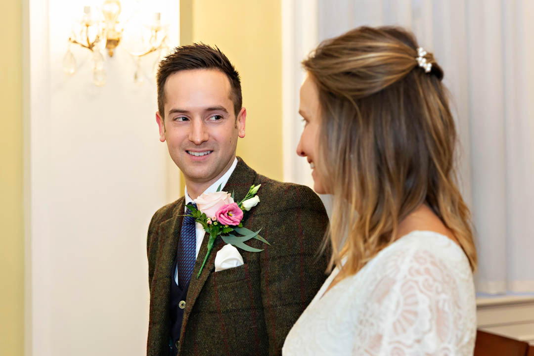 A groom in a winter jacket with pink buttonhole looks at his bride in a lace dress. Their wedding took place in Chelsea Old Town Hall's Rossetti Room.