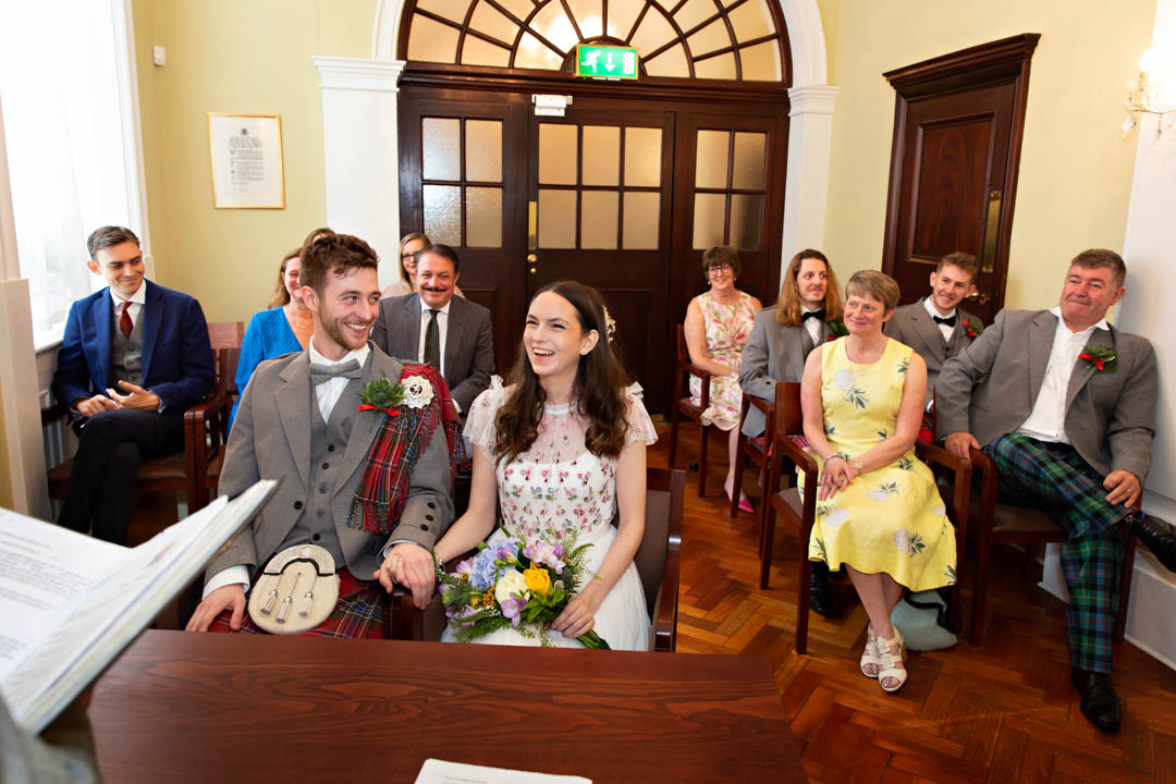 A Scottish groom and his bride in a flower-strewn dress hold hands during their Rossetti Room civil marriage ceremony. Their family and friends are looking on.