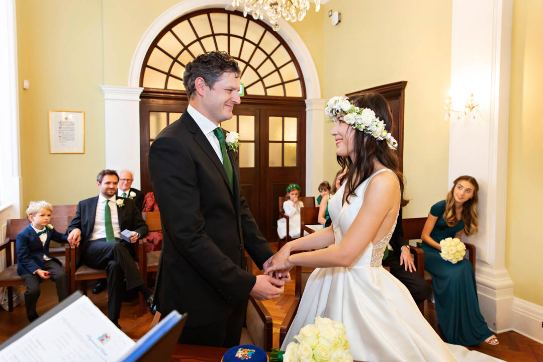 A bride wearing a green and white flower crown and full-length wedding dress holds the hands of her groom during their Rossetti Room wedding. Family and friends witness the moment.