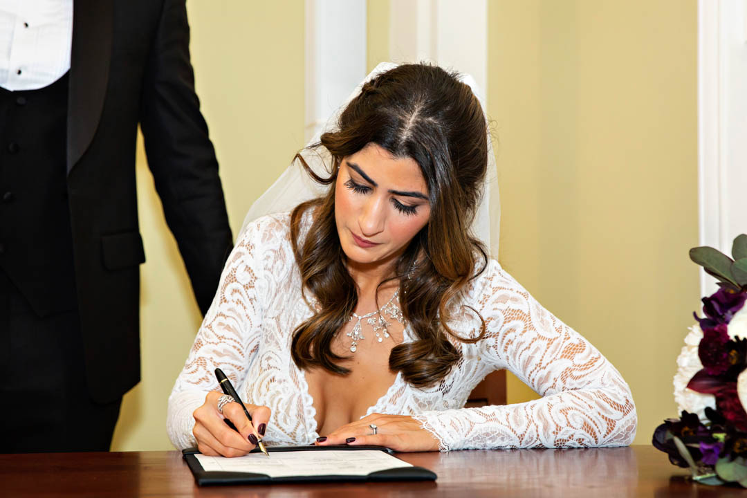 A bride in a lace wedding gown and veil signs the wedding paperwork during her civil marriage ceremony in the Rossetti Room.