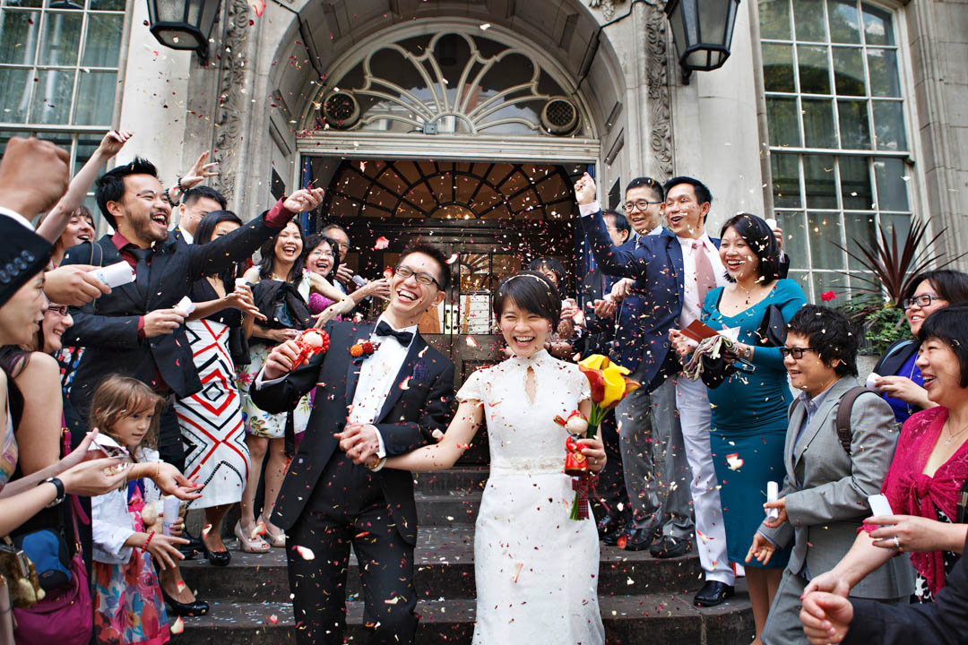 A Chinese bride and groom leave Chelsea Old Town Hall in a cloud of red rose petal confetti. The bride and groom are both carrying red figurines.