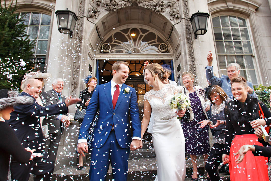 A bride in an ivory satin and lace wedding gown and her groom in a blue suit and red tie, walk down the steps of Chelsea Old Town Hall. Their guests are throwing a whirlwind of confetti.