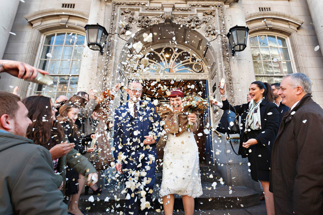 A red-headed bride and her groom can hardly be seen for a cloud of confetti. They're walking down the steps of Chelsea Old Town Hall on Kings Road.