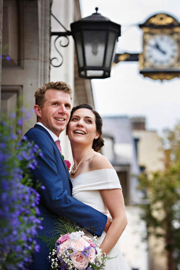 A bride in an off-the-shoulder white dress and a groom in a blue suit and pink tie stand cheek-to-cheek on the steps of Chelsea Old Town Hall. The hall's clock and a light are seen in the background.