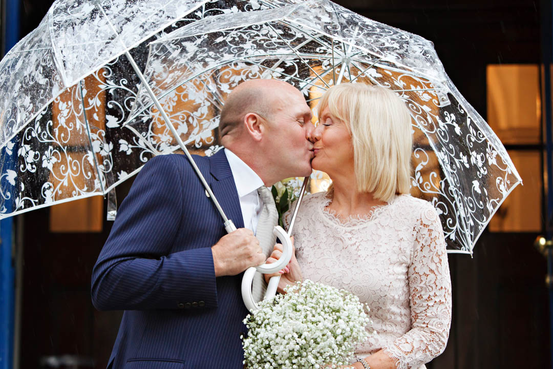 A bride in a pale lace dress who is holding a bouquet of gypsophila kisses her groom on the steps of Chelsea Old Town Hall. He's wearing a navy pin-striped suit. They're both holding umbrellas.
