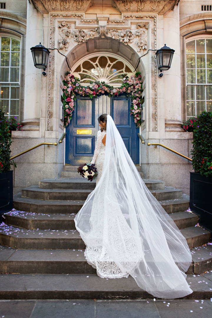 A bride in a white lace dress and a cathedral length veil stands on the steps of Chelsea Old Town Hall on Kings Road. The blue doors behind her are closed. They married in the Rossetti Room.