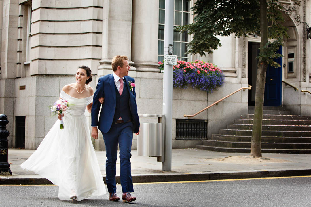 A bride in an off-the-shoulder full-length white gown and her groom in a blue suit and pink tie and buttonhole cross the Kings Road after their Rossetti Room civil marriage ceremony.