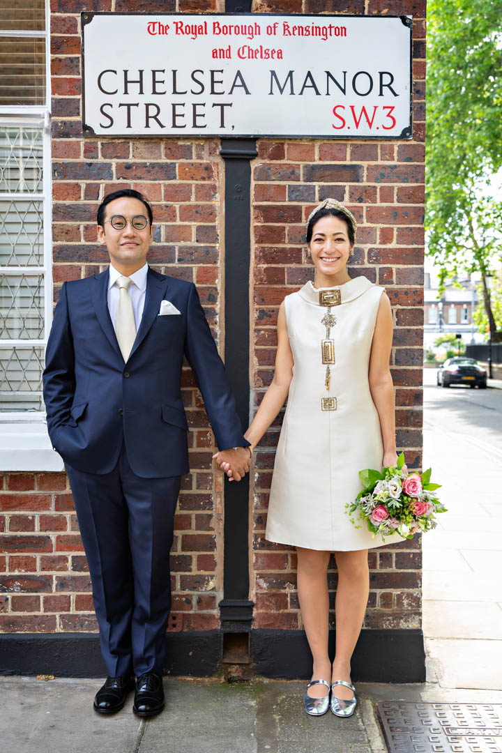 Newlyweds hold hands under the Chelsea Manor Street sign in London SW3. They married in the Rossetti Room, Chelsea Old Town Hall on the Kings Road, also in the Royal Borough of Kensington and Chelsea.