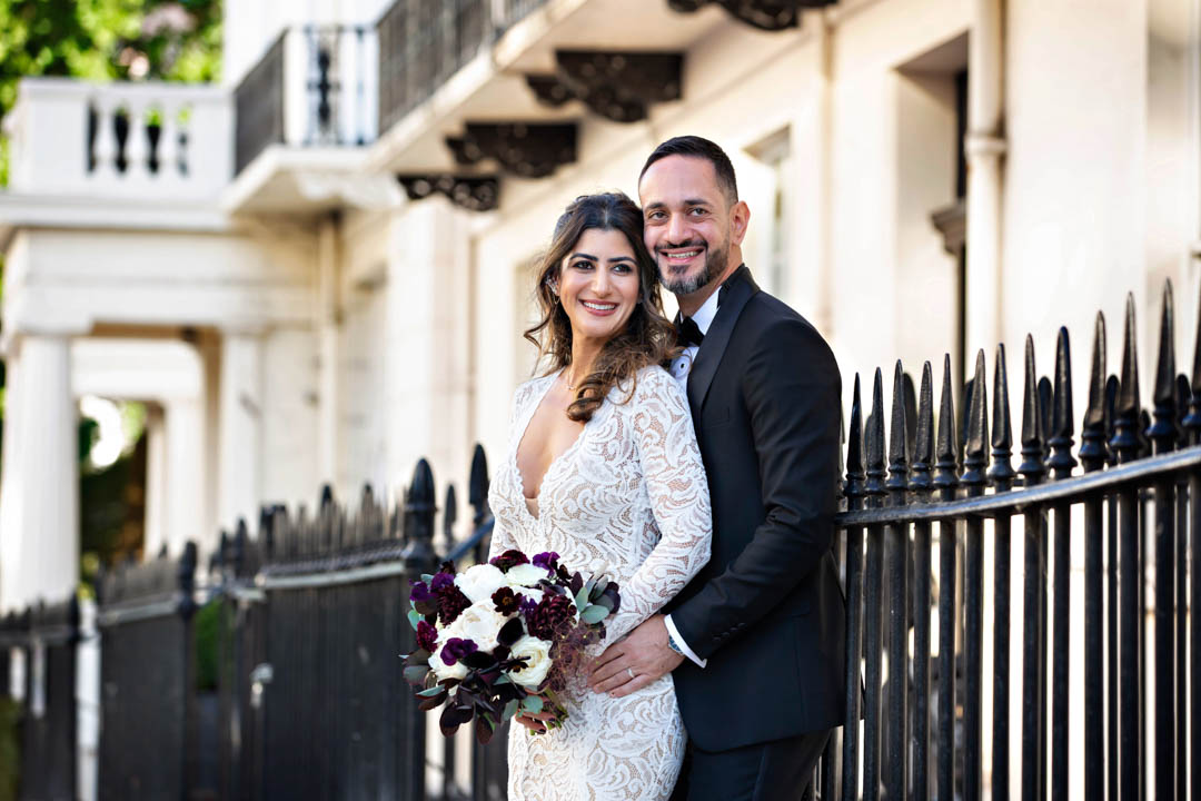 A bride in a fitted white lace dress and holding a white rose and deep maroon bouquet leans against her groom in evening dress. They're both standing near black metal railings in London's Royal Borough of Kensington and Chelsea, after being married in the Rossetti Room at Chelsea Old Town Hall.