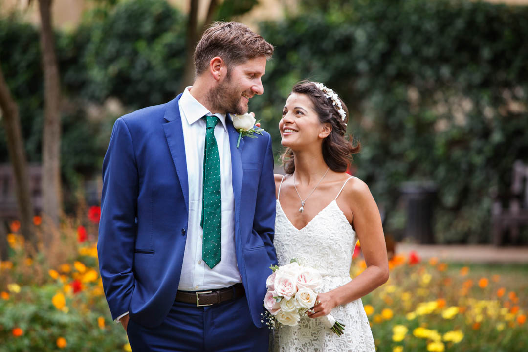 A newlywed couple walk through a colourful garden after their Chelsea Old Town Hall wedding in the Rossetti Room. A bride in a textured white dress with shoestring straps and wearing a white hair band looks at her new husband. The man is wearing a bright blue suit with a green tie and white rose buttonhole. They're walking through gardens with yellow, orange and red flowers. The bride is holding a pink and white rose bouquet.