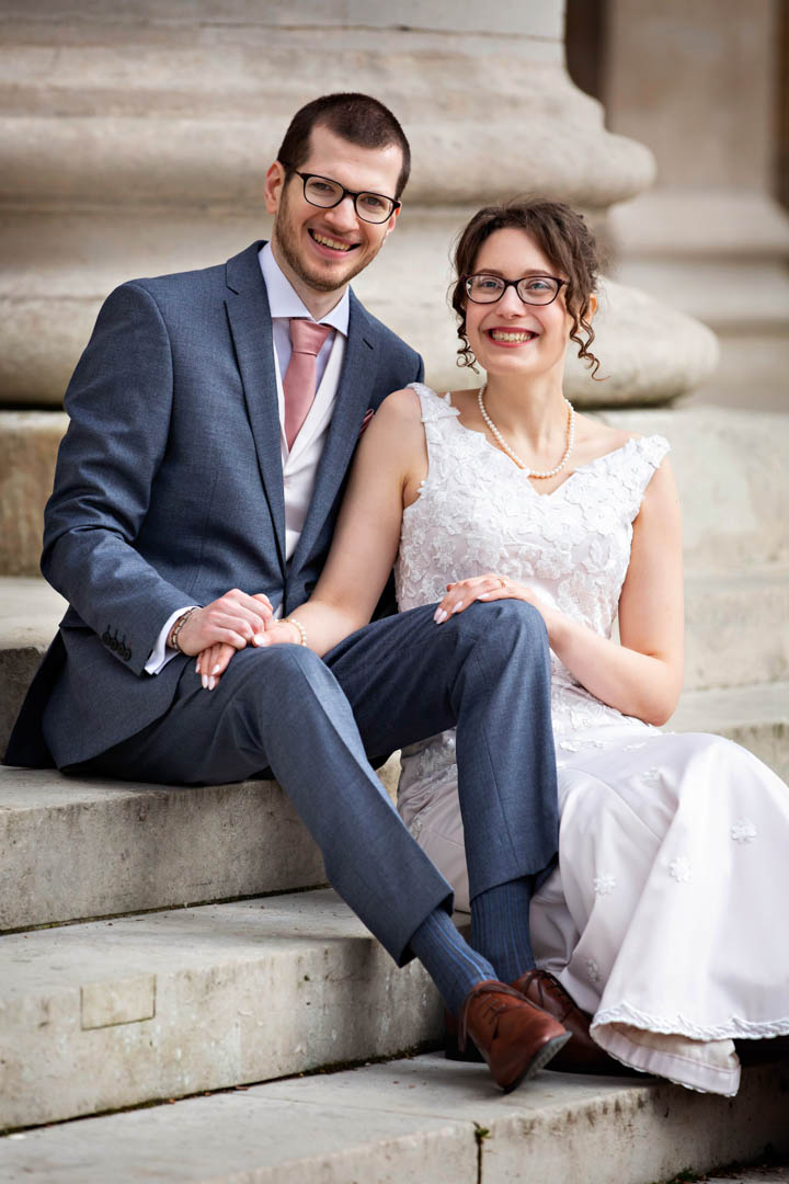 A bride wearing a white, sleeveless dress sits on some stone steps in London's Chelsea, her hands resting on the groom's knees. He is wearing a pale grey suit, cream waistcoat and pink tie.