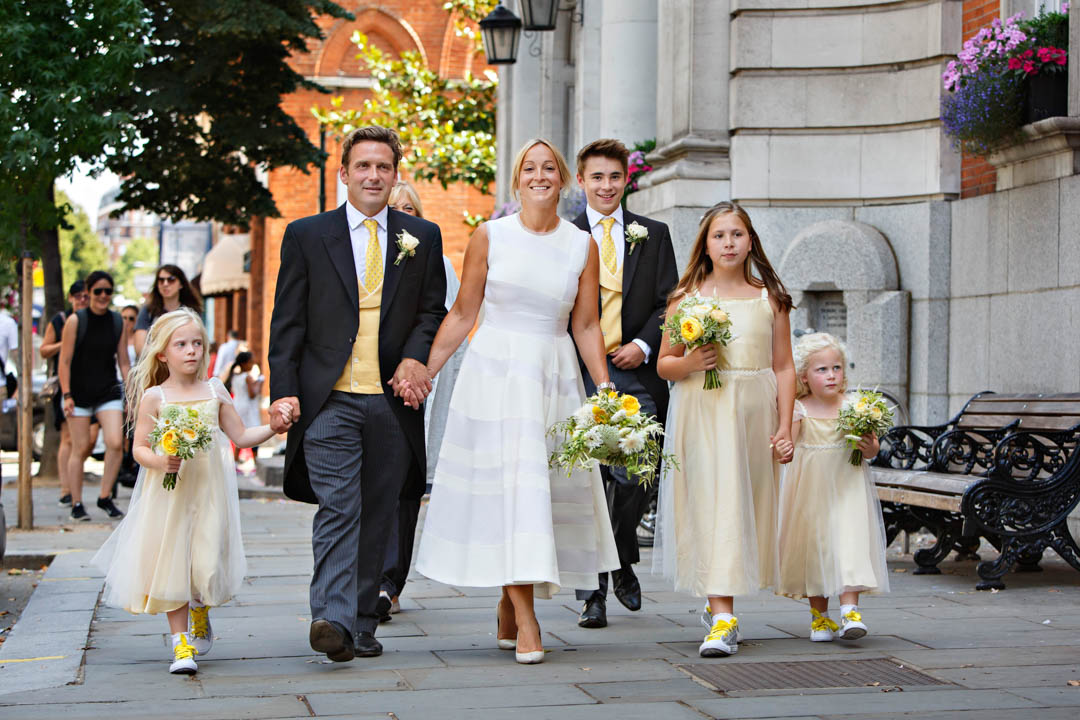 A bride in a midi white dress walks down Kings Road with her husband and their children dressed in yellow and carrying yellow bouquets. The men have yellow ties, pale yellow waistcoats and are wearing morning suits. They had their wedding at Chelsea Old Town Hall's Rossetti Room.