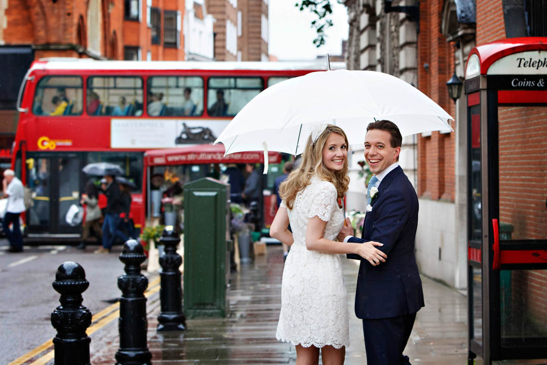 A bride in a short white lace dress and groom in a blue suit and tie stand under a white umbrella. There is a red London bus in the background.