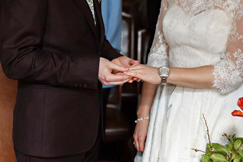 A close-up photograph of the groom placing the wedding ring on the bride's finger as they exchange rings during their intimate wedding.