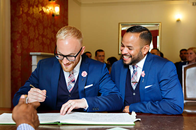 Two grooms wearing matching blue suits are full of laugher as they sign the register during their wedding at Southwark Register Office.