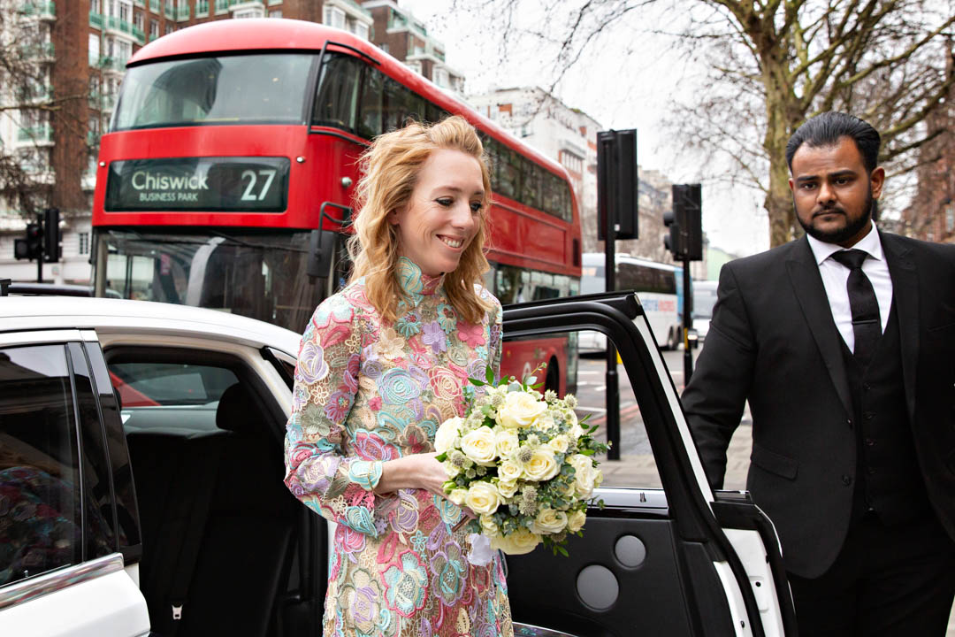 A bride in a pastel florals dress arrives for her wedding ceremony with a red London bus in the background