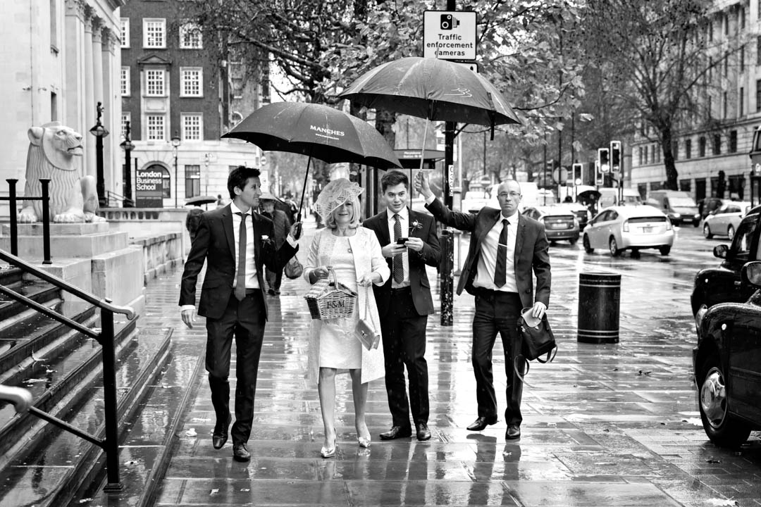 The mother of the groom arrives at The Old Marylebone Town Hall in the pouring rain, as the groomsmen protect her with two umbrellas.