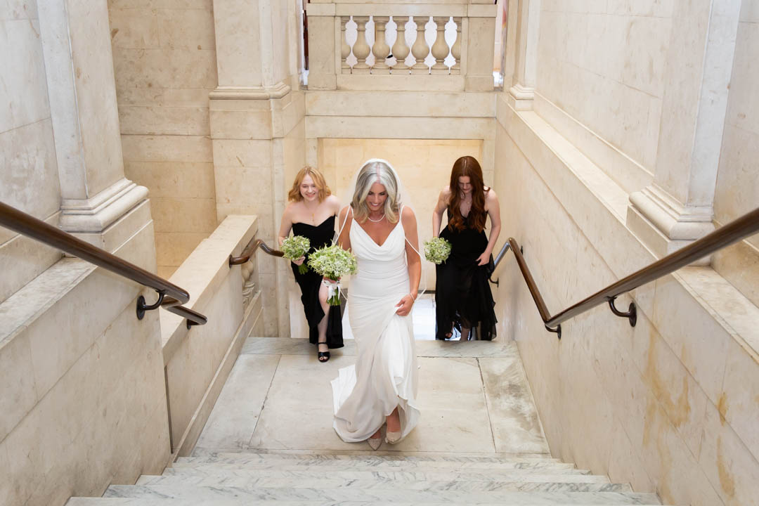 A bride and her two bridesmaids walk up the stairs of The Old Marylebone Town Hall to enter her wedding ceremony.
