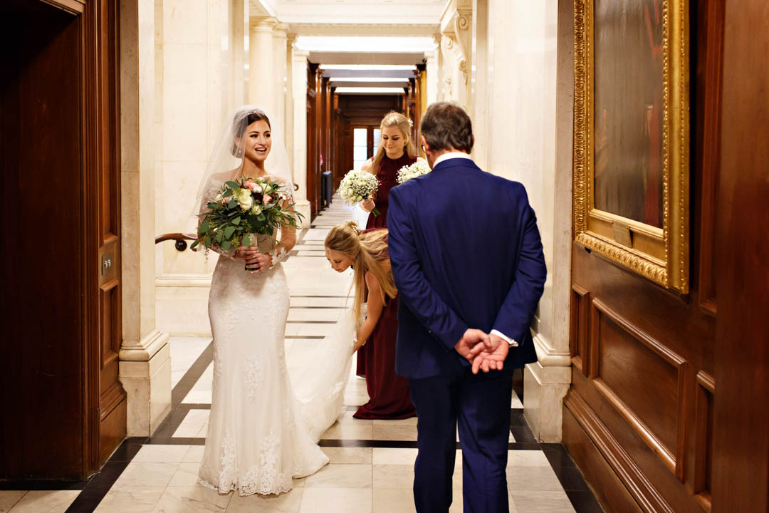 The bride walks down the corridor of the Old Marylebone Town Hall while the bridesmaids make final adjustments to the train.