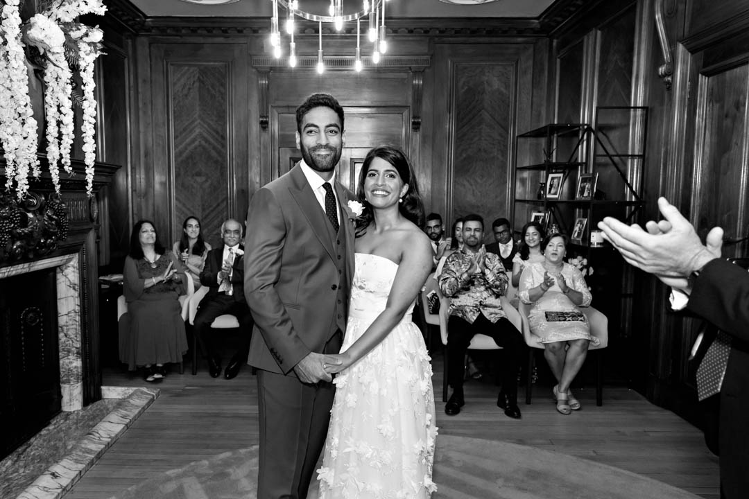 A black and white photograph of a bride and groom celebrating the end of their wedding ceremony as their wedding guests applaud.